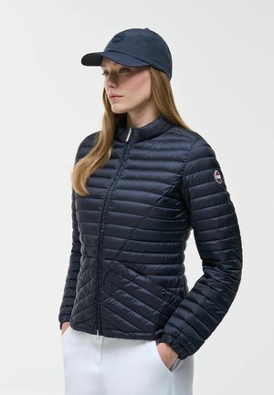 Ženska z dolgimi lasmi, oblečena v temnomodro prešito jakno, ustrezno baseball kapo in bele hlače, stoji z rokami v žepih in gleda levo.