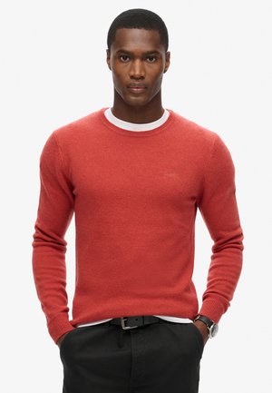 Rød strikket sweater med rund hals, der har lange ærmer og en tekstureret overflade. Båret over en hvid skjorte.