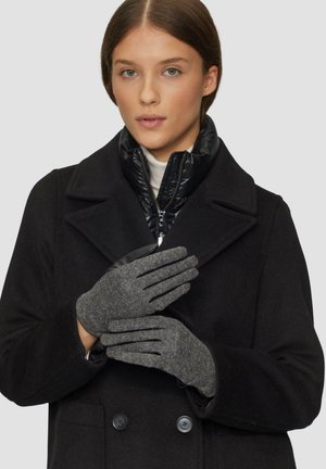 Frau, die graue Wollhandschuhe und einen schwarz-gestreiften Doppelreiher mit einem Reißverschlusskragen darunter trägt, posiert mit verschränkten Händen.