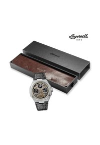 Zilveren horloge met een skeleton wijzerplaat, zwarte leren band en gouden accenten in een zwarte doos met bruine leren binnenkant.