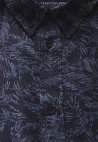 Námořnická modrá košile s krátkým rukávem a texturovanou tkaninou, s listovým vzorem v světlejší modré barvě, dvěma černými knoflíky a špičatým límcem.