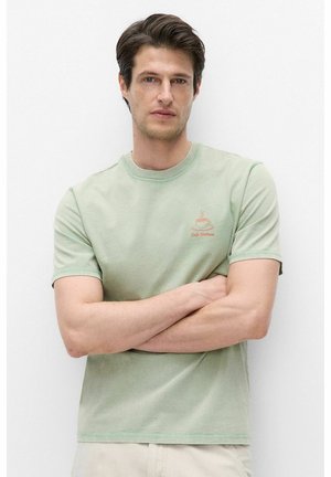 Young man with short brown hair wearing a light green T-shirt with a coffee cup logo, standing with arms crossed against a white background.