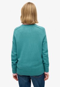 Lyshåret person iført en teal strikket sweater over en lyserød skjorte og blå jeans, stående med ryggen mod hvid baggrund.