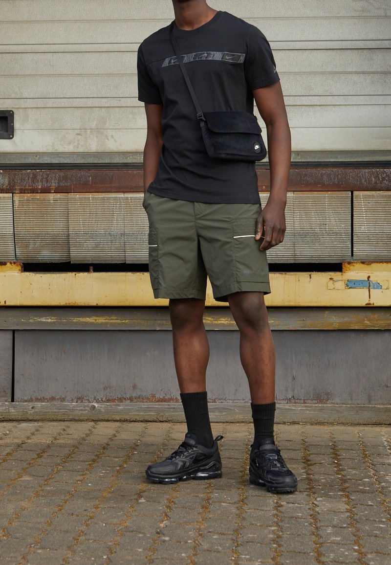 Homme portant un t-shirt noir Nike, un sac bandoulière noir, un short cargo olive, des chaussettes noires et des baskets noires, debout sur un sol pavé.