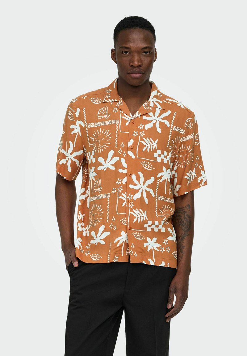 Hombre vistiendo una camisa naranja de manga corta con botones y patrones abstractos en blanco de sol, hojas y ojos, combinada con pantalones negros.