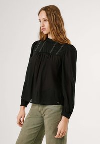 Zwarte blouse met lange mouwen en een hoge hals, met geplooide details en witte stikselaccenten. Gemaakt van lichtgewicht stof met een zachte textuur.