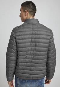 Lühikeste juustega mees, kes kannab halli toppkattega jopet ja siniseid teksaseid, seisab seljaga, taustaks lihtne hall sein.