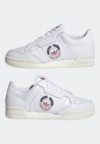 Sneaker bianche in pelle con superficie testurizzata, caratterizzate da logo e dettagli ricamati in rosso e nero, insieme a una punta perforata e lacci bianchi.