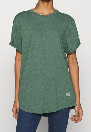 Person iført en løs grøn kortærmet t-shirt med opbukne ærmer og blå jeans, stående mod en ensfarvet baggrund.