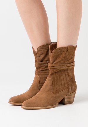 Hnědé semišové kotníkové boty s volným tvarem, špičatou špičkou, perforovanými detaily a podpatkem se strukturou.