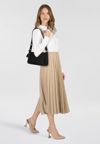 Bolso de cuero negro con correa ajustable, llevado sobre una camisa blanca abotonada. Falda plisada beige y zapatos de tacón alto beige completan el conjunto.
