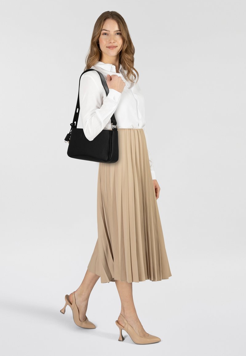 Bolso de cuero negro con correa ajustable, llevado sobre una camisa blanca abotonada. Falda plisada beige y zapatos de tacón alto beige completan el conjunto.