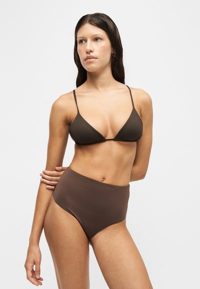 Femme aux longs cheveux foncés portant un haut de bikini triangle marron foncé et un bas de bikini taille haute, debout avec un bras croisé dans le dos.