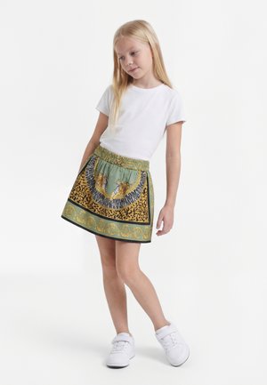 Jeune fille en t-shirt blanc, jupe verte à motifs tigre et léopard, et baskets blanches, debout sur fond blanc.