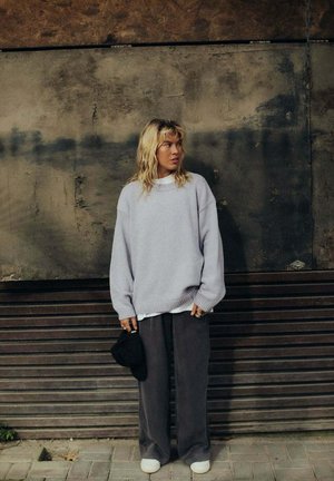 Za duży, jasnofioletowy, dziergany sweter z żebrowanymi mankietami, zestawiony z luźnymi szarymi spodniami i białymi trampkami. Nosi czarną torbę.