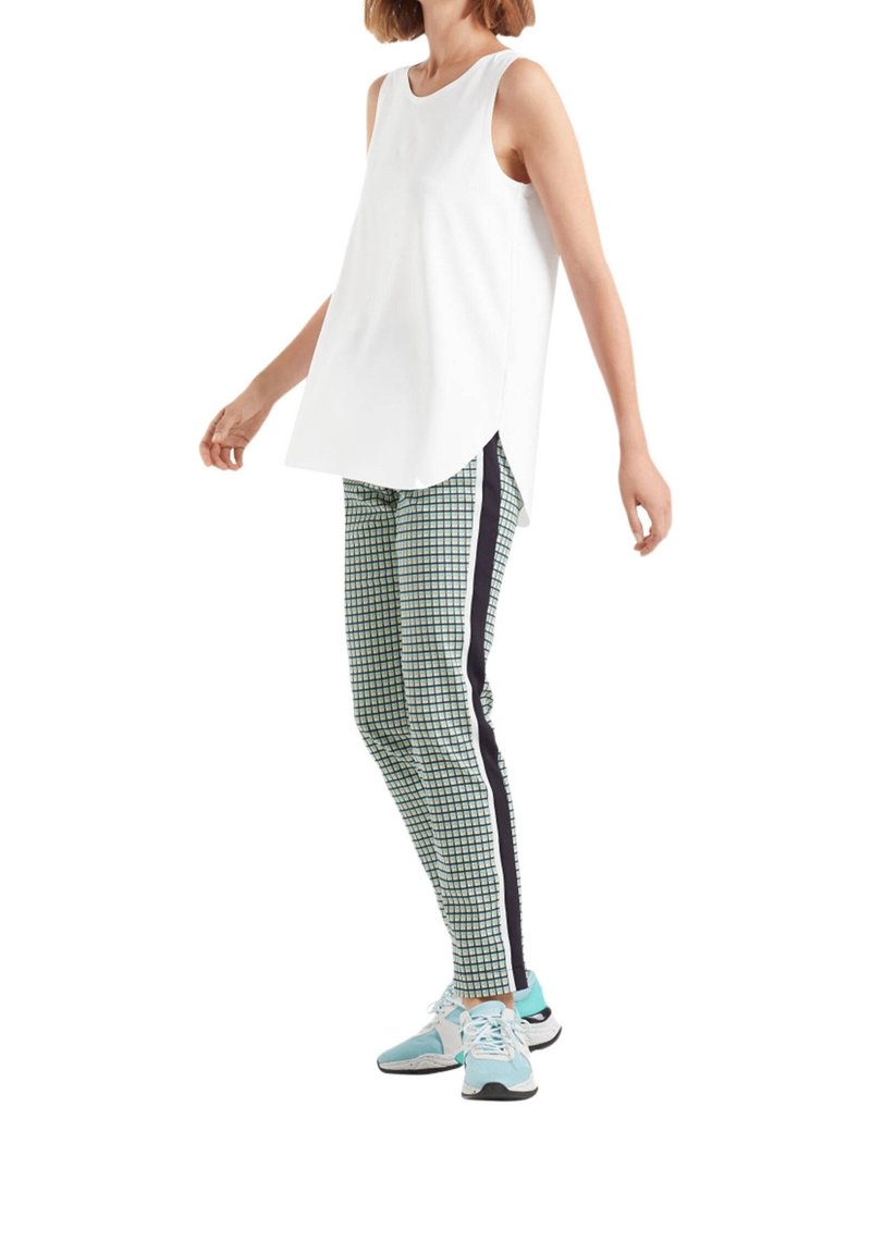 Haut blanc sans manches avec ourlet incurvé, associé à des leggings verts à motifs avec des bandes latérales noires et des chaussures de sport bleues.