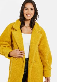 Abrigo grande, texturizado y de color amarillo brillante, con solapas en pico, botones dobles y hombros caídos, llevado sobre una blusa blanca.