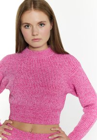 Maglione corto rosa con un collo alto, texture a coste e maniche lunghe, che mostra una combinazione di fibre rosa chiare e scure.