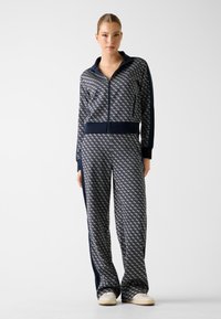 Granatowy dres z diamentowym wzorem, kurtka na zamek i elastycznym pasem. Miękki materiał z kontrastowymi paskami po bokach i ściągaczami na rękawach. Wygodne, luźne dopasowanie.