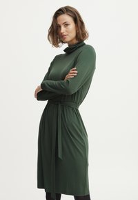 Žalia ilgomis rankovėmis suknelė, pagaminta iš minkštos, elastingos medžiagos. Yra aukštai sukurtas apykaklės iškirpimas ir diržas, su kuriuo galima suformuoti siluetą.