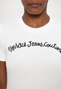 Balta kokvilnas T-krekls ar apaļu kakla izgriezumu, uz kura ir melni izšūti vārdi "Versace Jeans Couture" izliekta dizainā.