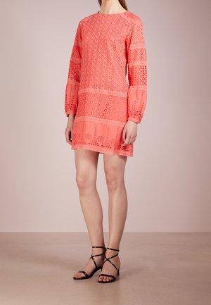 Femme portant une robe en dentelle corail rose à manches longues avec des découpes florales et des sandales noires à talons bas et à lanières, debout devant un fond uni.