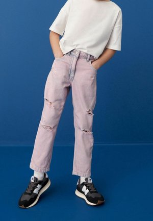 Enfant portant un t-shirt blanc, un jean rose clair déchiré, des chaussettes blanches et des baskets noir et blanc, debout devant un fond bleu.