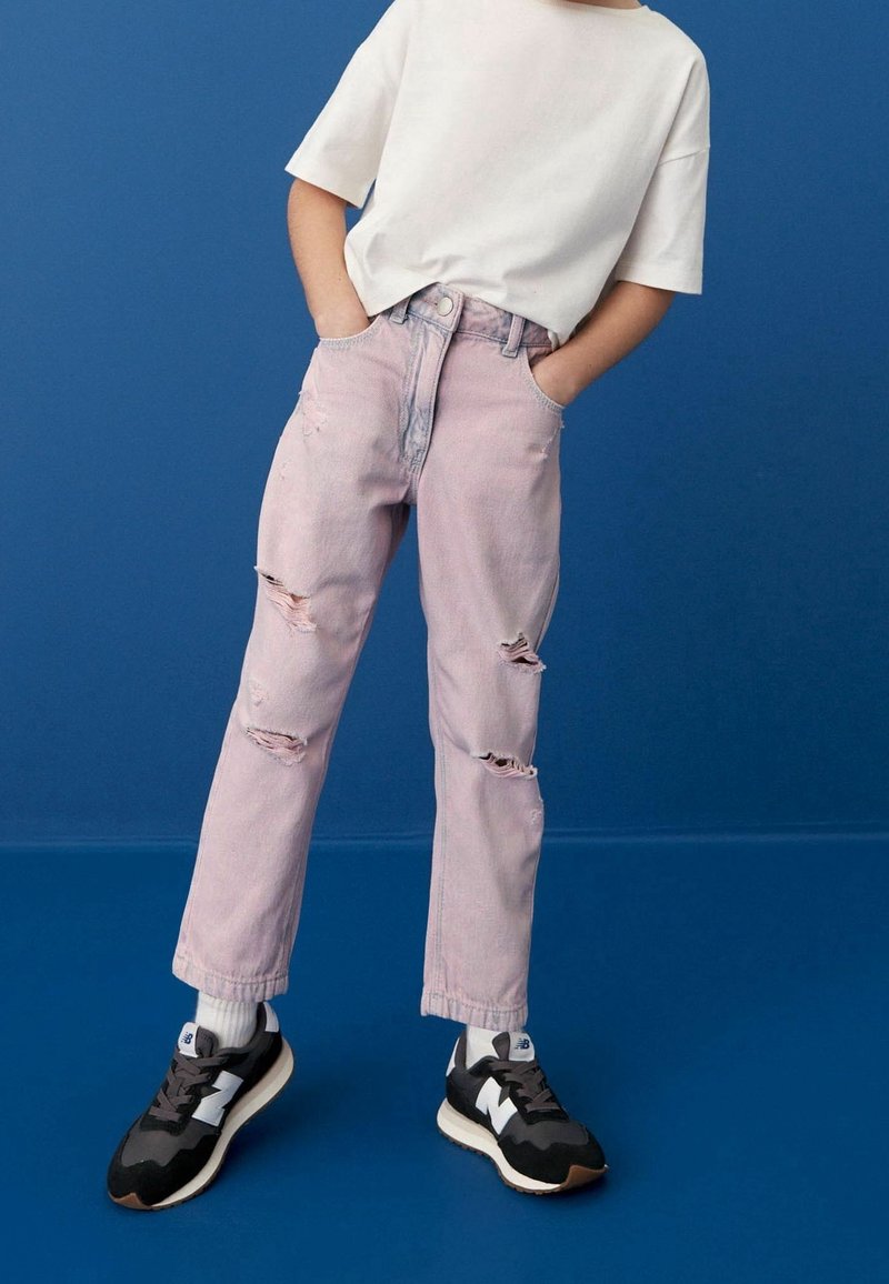 Enfant portant un t-shirt blanc, un jean rose clair déchiré, des chaussettes blanches et des baskets noir et blanc, debout devant un fond bleu.