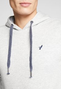 Ljust grå hoodie med dragsko och marinblå accenter. Innehåller en liten broderad fågel-logotyp på bröstet; tillverkad av ett mjukt material.