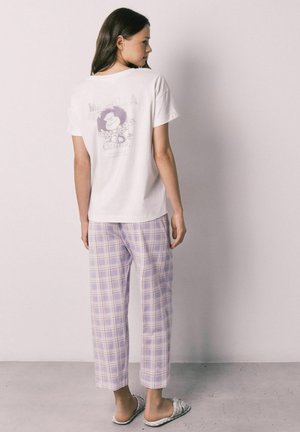 Femme de dos portant un T-shirt blanc avec un dessin animé et un pantalon à carreaux violet clair, debout contre un mur blanc uni.