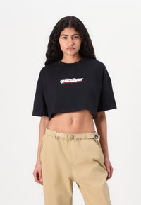 T-shirt nera corta in cotone, con un logo bianco e rosso sul davanti. Abbinata a pantaloni beige chiaro e larghi.