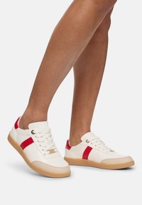 Sneakers bianchi con strisce rosse, design con lacci e dettagli dorati. Presentano una suola in gomma e una texture liscia.