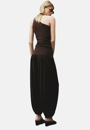 Dark brown jumpsuit with a one-shoulder design, ruched texture, and loose, tapered legs. Paired with black shoes and sunglasses.