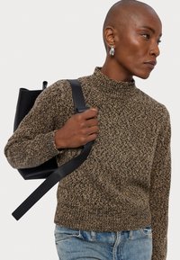 Un pull texturé marron et noir avec un col haut, associé à un sac à dos noir. La personne a les cheveux courts et tient la sangle du sac.