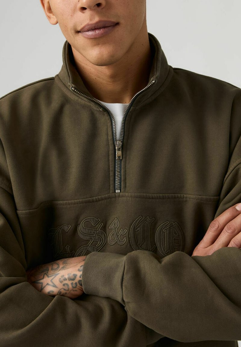 Polaire verte olive à fermeture éclair quart, avec logo brodé en ton sur ton. Comprend un col montant et une texture douce et confortable.