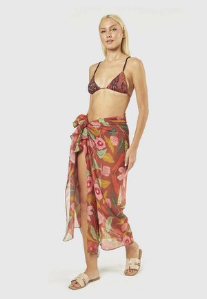 Donna con capelli lunghi e biondi che indossa un top bikini marrone a fantasia, una gonna a portafoglio floreale legata in vita e sandali beige scivolati.