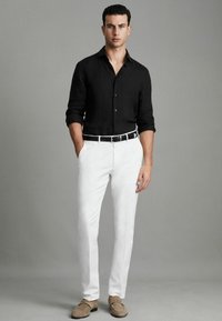 Schwarze Leinenbluse, weiße maßgeschneiderte Hose, schwarzer Gürtel und beigefarbene Slipper. Das Model steht mit den Händen in den Taschen vor einem grauen Hintergrund.