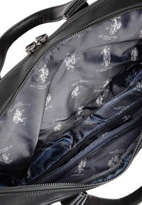 Interior de una bolsa de cuero negro con textura suave, compartimentos con cremallera y un forro azul marino con patrones repetidos del logo en gris claro.