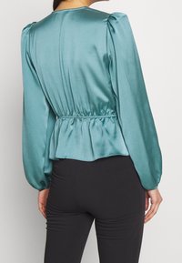 Blouse en satin sarcelle avec manches longues bouffantes, froncée à la taille, et une texture lisse et brillante. Le dos présente un col arrondi et un ourlet à basque.