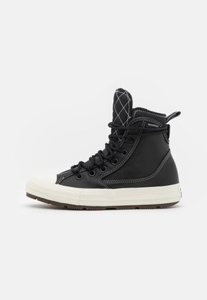 Bottine de sneaker haute imperméable noire avec semelle en caoutchouc blanche, coutures contrastées, languette matelassée et lacets noirs, vue de profil.