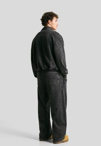 Uomo con giacca di denim nera larga e pantaloni, indossa stivali beige, in piedi di lato su uno sfondo grigio uniforme.