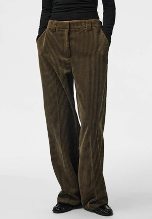 Personne portant un pantalon en velours côtelé marron ample, les mains dans les poches et des chaussures noires, debout devant un fond clair uni.