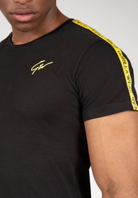 Zwarte katoenen T-shirt met een gele logo en schouderbanden met de tekst "GORILLA WEAR". Regular fit, korte mouwen, gladde textuur.