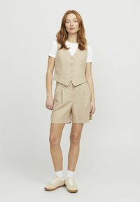 Chaleco sin mangas beige con cierre de botones, combinado con pantalones cortos a juego. Lleva una camiseta blanca debajo y se completa con zapatillas crema.