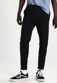 Pantalon noir ajusté à coupe slim avec une fermeture à bouton, jambes fuselées et détails zippés aux chevilles, assorti à des baskets noires et des chaussettes blanches à la cheville.