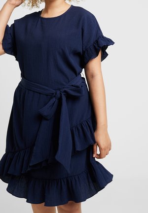 Kvinde iført en marineblå kjole med korte ærmer med flæser på ærmer og kant, bundet i taljen med et bælte, stående foran en ensfarvet baggrund.