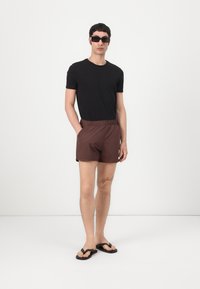 Svart T-skjorte, brune badebukser med elastisk liv, og svarte flip-flopper. Modellen bruker firkantede solbriller. Enkel og uformell stil.