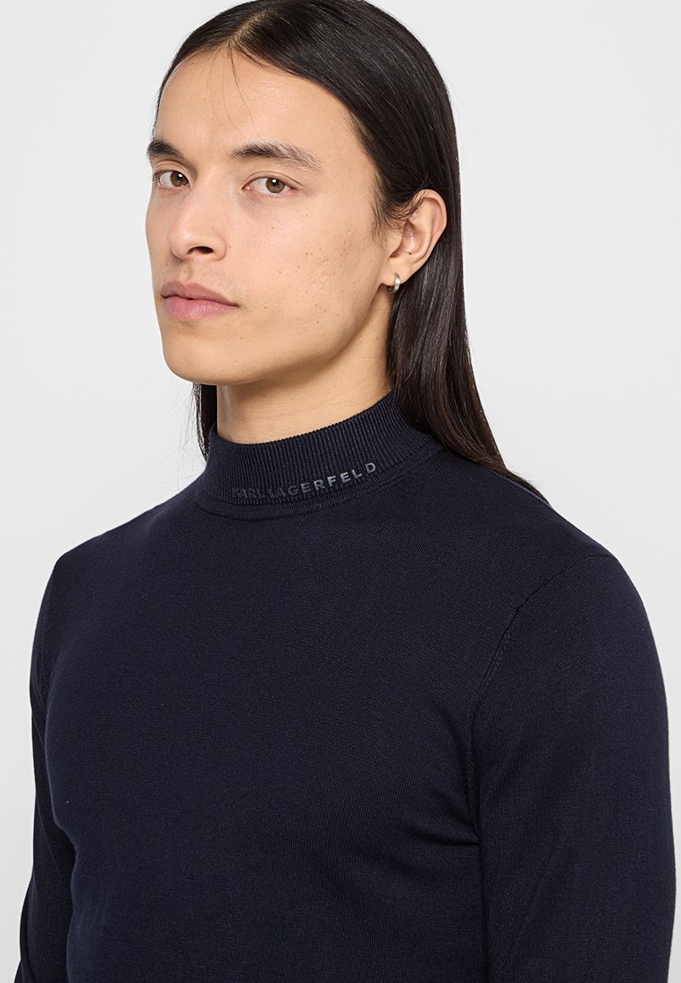 Marineblauer, taillierter Rollkragenpullover mit geripptem Kragen, mit einem Logodetail in der Nähe des Kragens, präsentiert an einem Modell mit langen Haaren.