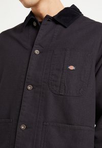 Giacca nera in tela con colletto in corduroy, bottoni a scatto e una tasca sul petto con un logo del marchio multicolore. Texture liscia.