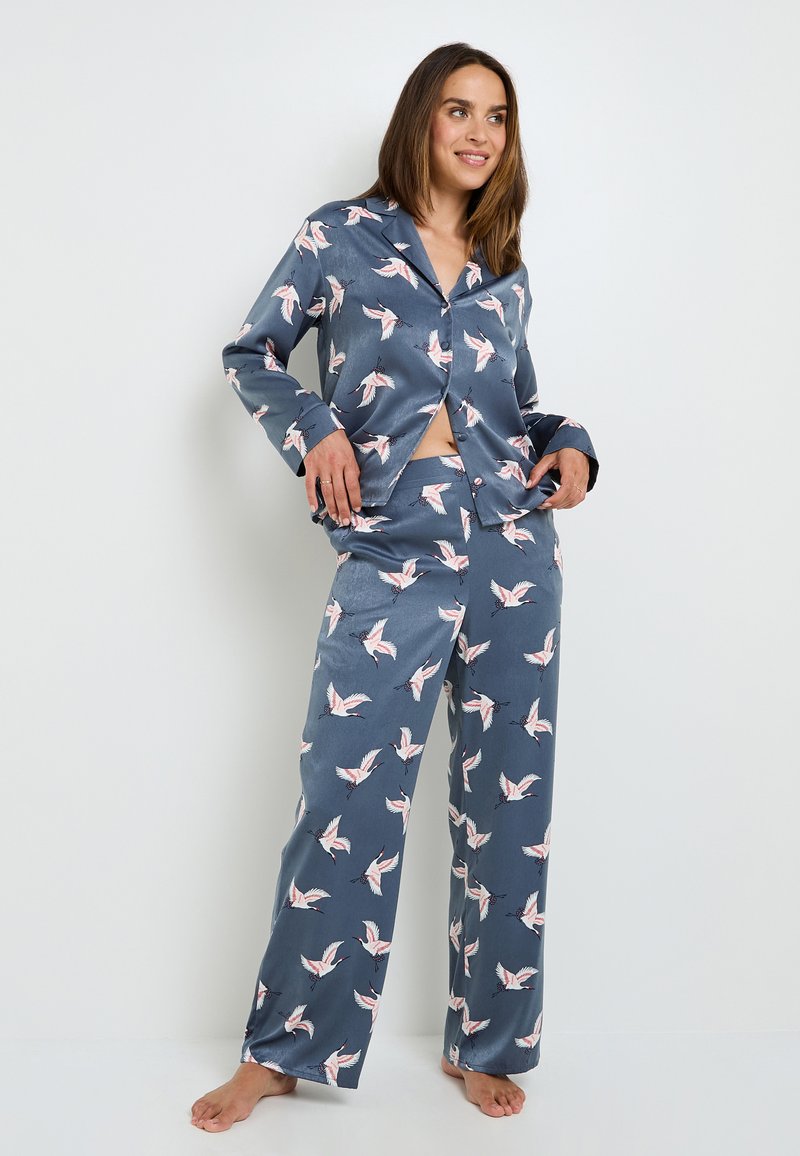 Ensemble de pyjama en soie avec un fond bleu-gris et un imprimé de grues blanches et roses. Le haut a un col et une fermeture à boutons ; le pantalon est ample.